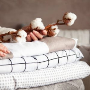 istockphoto-1279913217-612×612 Well groomed woman hands holding the cotton branch with pile of neatly folded bed sheets, blankets and towels. Production of natural textile fibers. Manufacture. Organic product.